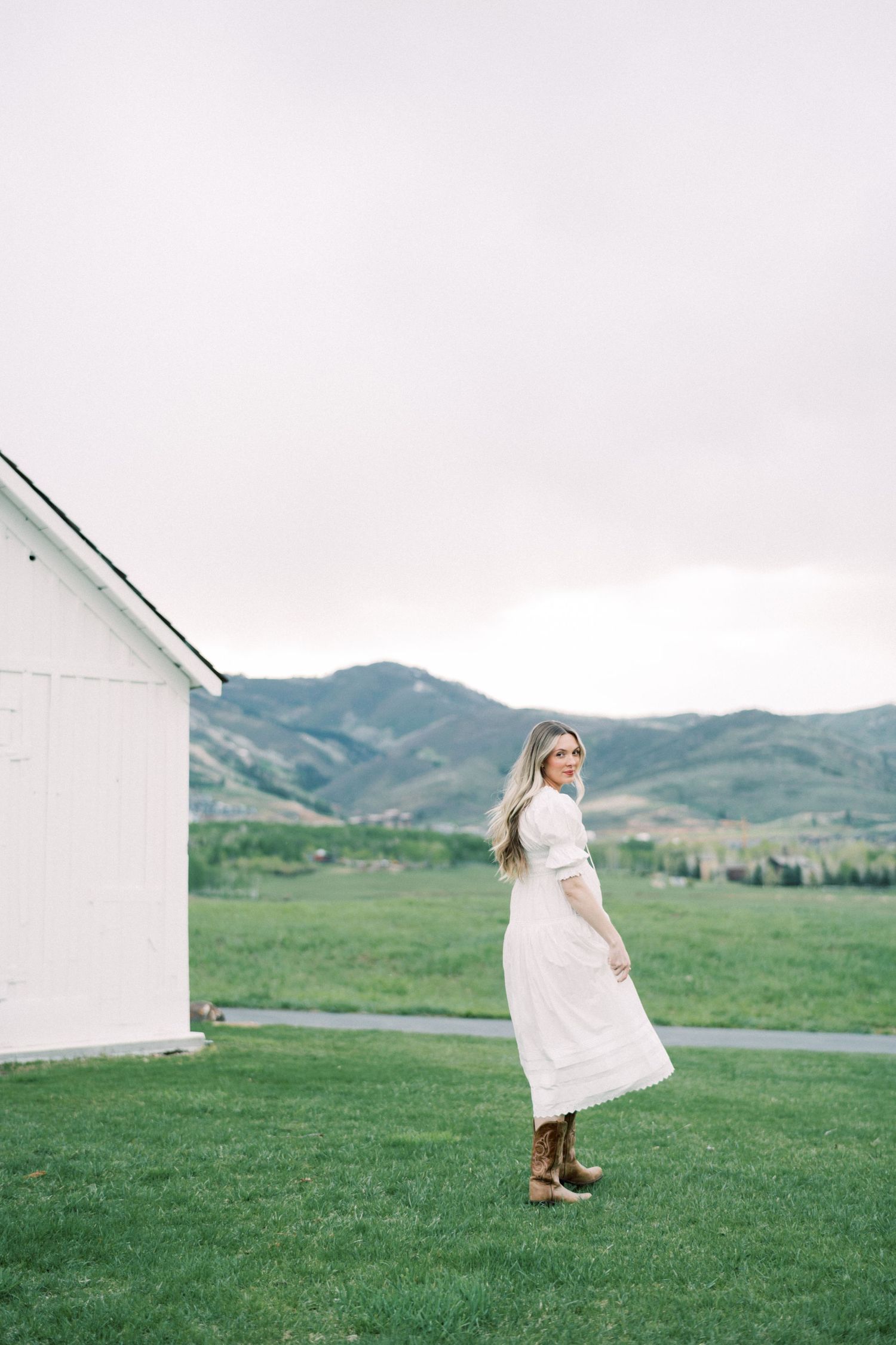 Figure in a flowing white dress stands on green grass with mountains and a white building in the background.