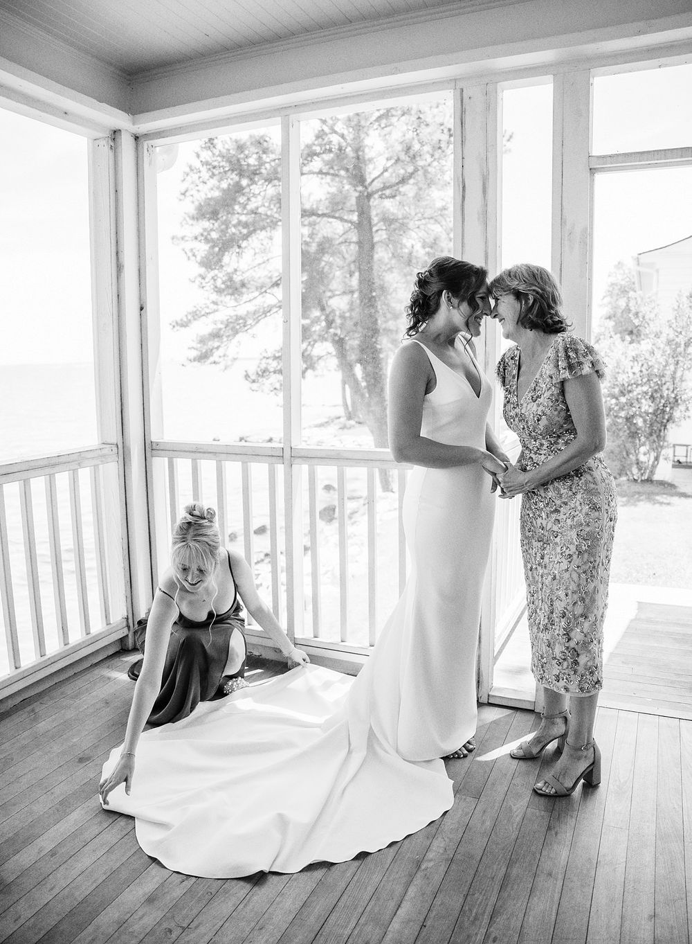 Black and white photograph of two people in elegant dresses sharing an intimate moment on a sunlit porch.