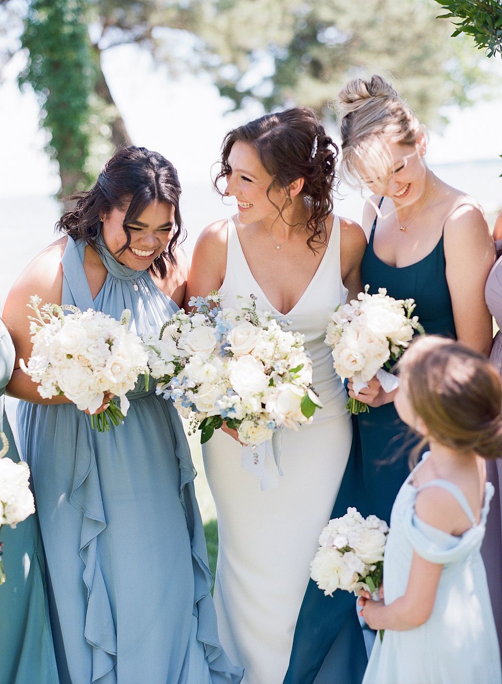 Bridesmaids in elegant blue gowns hold stunning white floral bouquets at an outdoor wedding celebration.