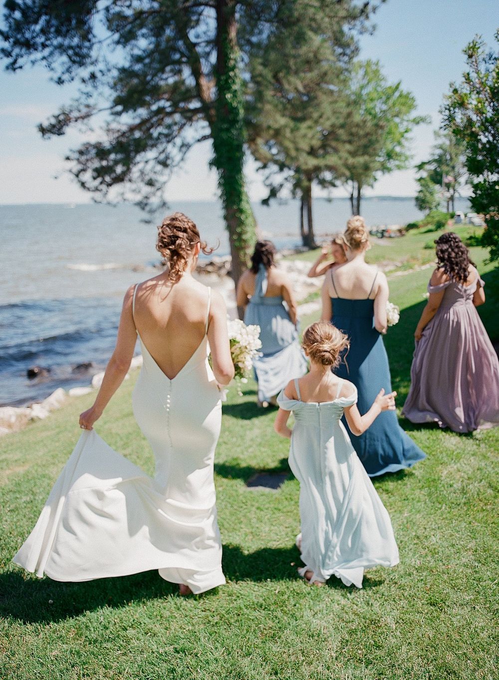 Wedding party walking along grassy waterfront area with trees on a sunny summer day.