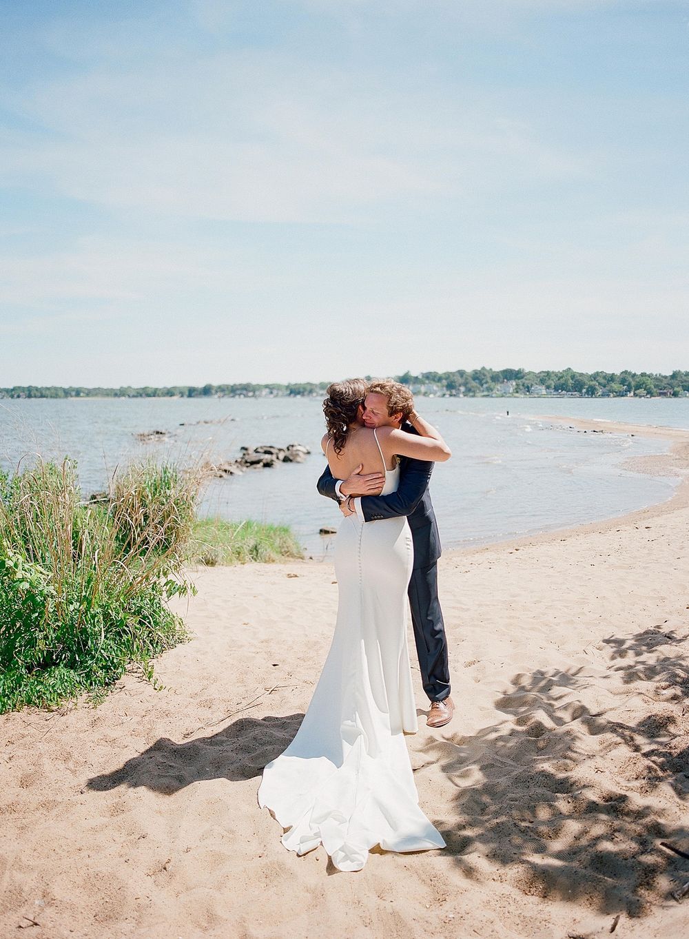 A couple embraces on a sandy beach with a lake and natural shoreline in the background under blue summer skies.
