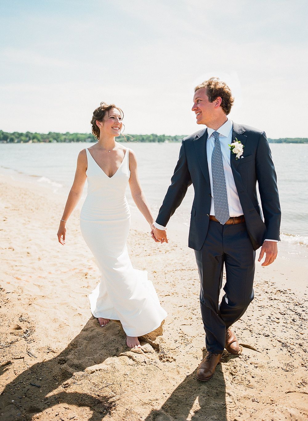 Couple in wedding attire walking and laughing together on a sunny beach with water in the background.