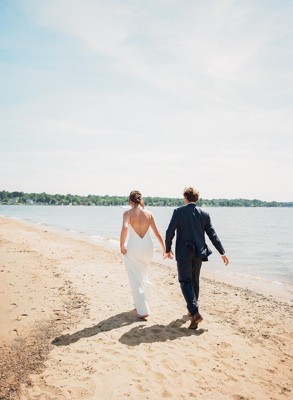 A couple in wedding attire walks hand in hand along a sandy beach with blue water and trees in the background.