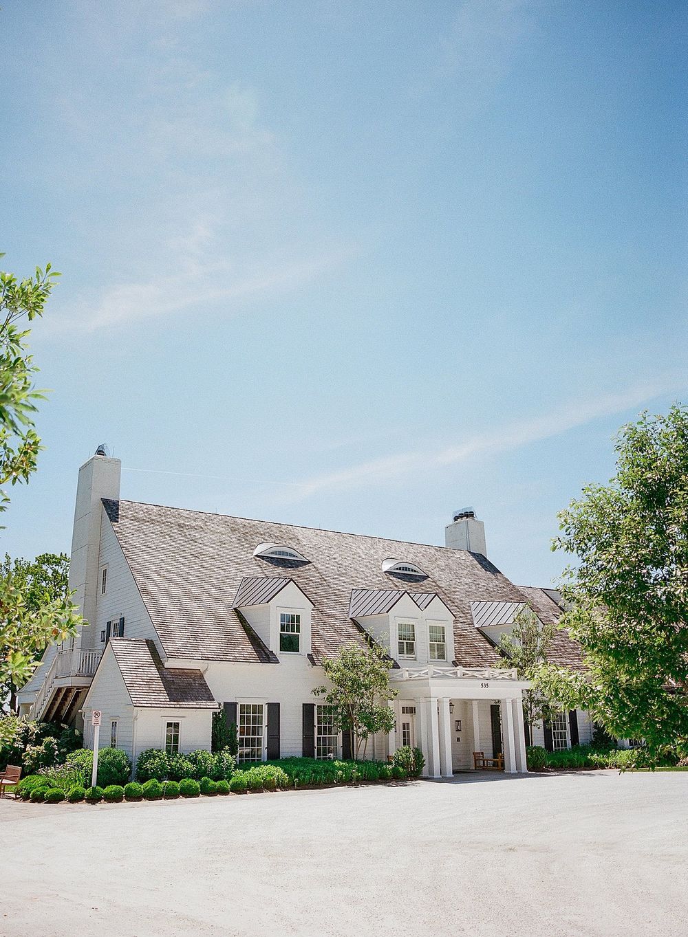 Beautiful country estate home with thatched roof, dormers, and white exterior surrounded by trees on a sunny day.