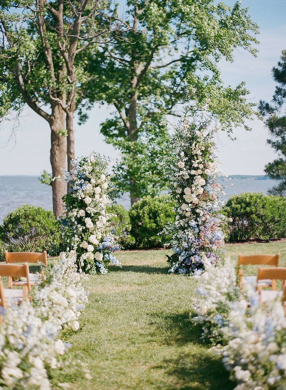 Romantic outdoor wedding ceremony setup with floral arch and wooden chairs overlooking a waterfront under tall trees.