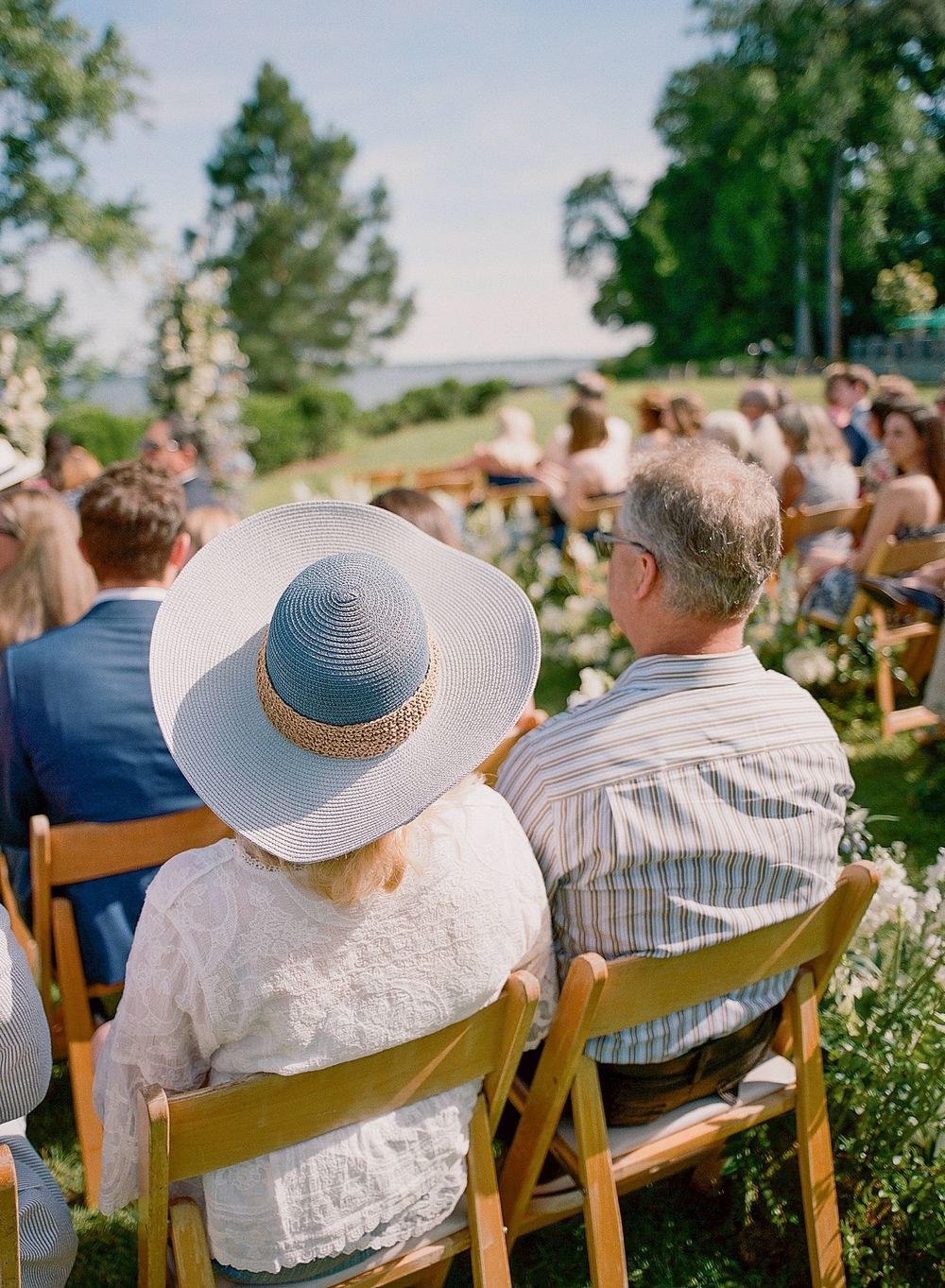 Guests seated in wooden folding chairs watch an outdoor ceremony against a scenic natural backdrop.