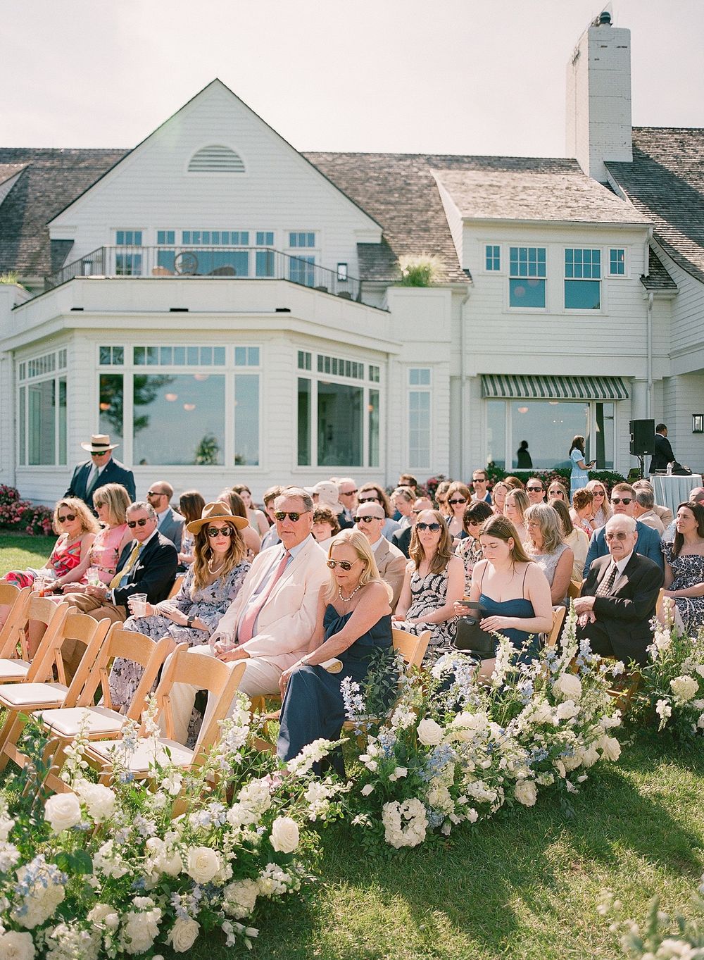 Outdoor wedding ceremony setup with wooden chairs and white floral arrangements in front of an elegant coastal-style house.