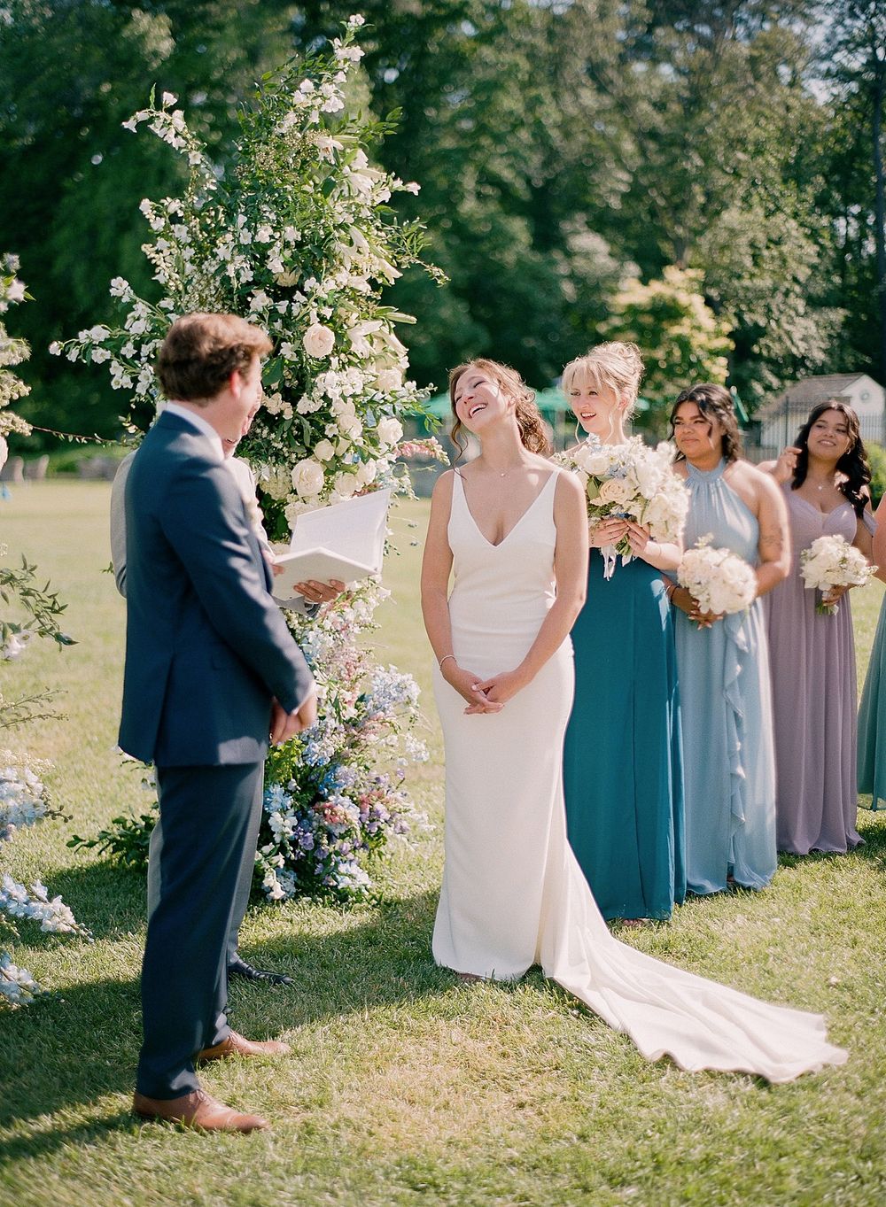 An outdoor wedding ceremony with a floral arch and bridesmaids in varying shades of blue and purple dresses.