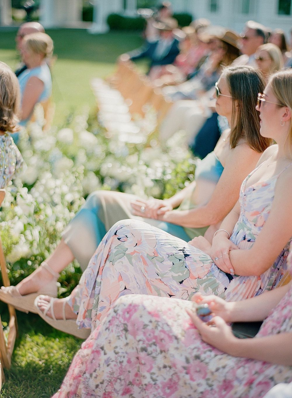 People in floral summer dresses sit outdoors on the grass at a garden party with white flowers blooming nearby.