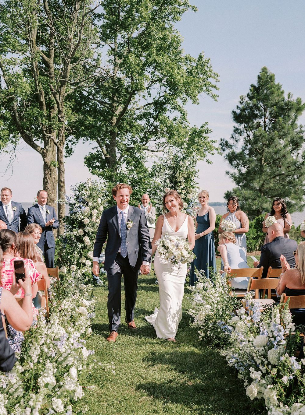 Outdoor wedding ceremony among flowering trees and delicate white floral arrangements lining a grassy aisle.