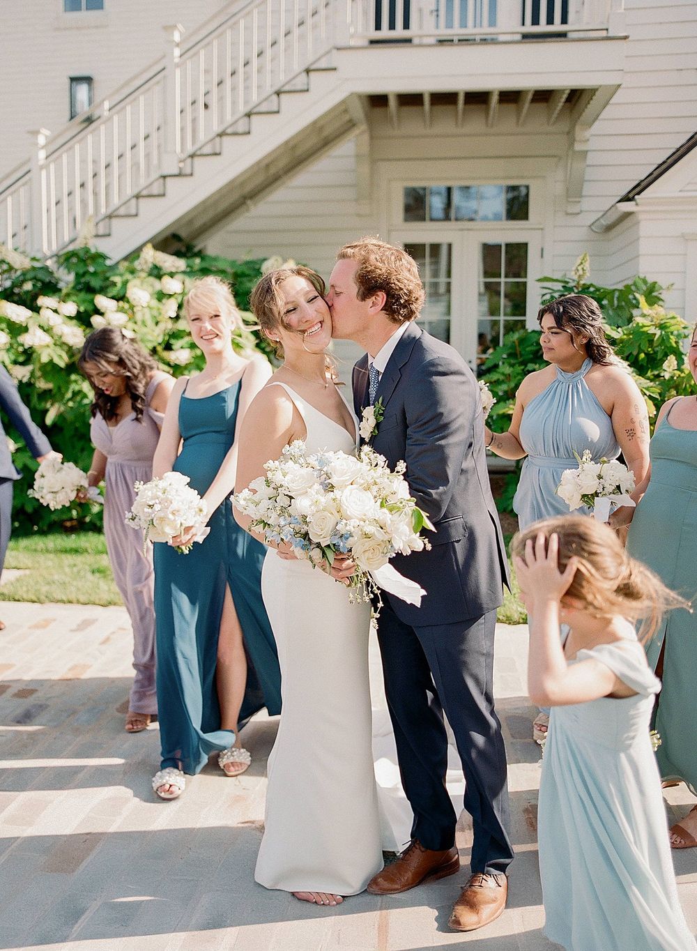 A wedding party poses on stone steps surrounded by hydrangeas with bridesmaids in blue dresses.