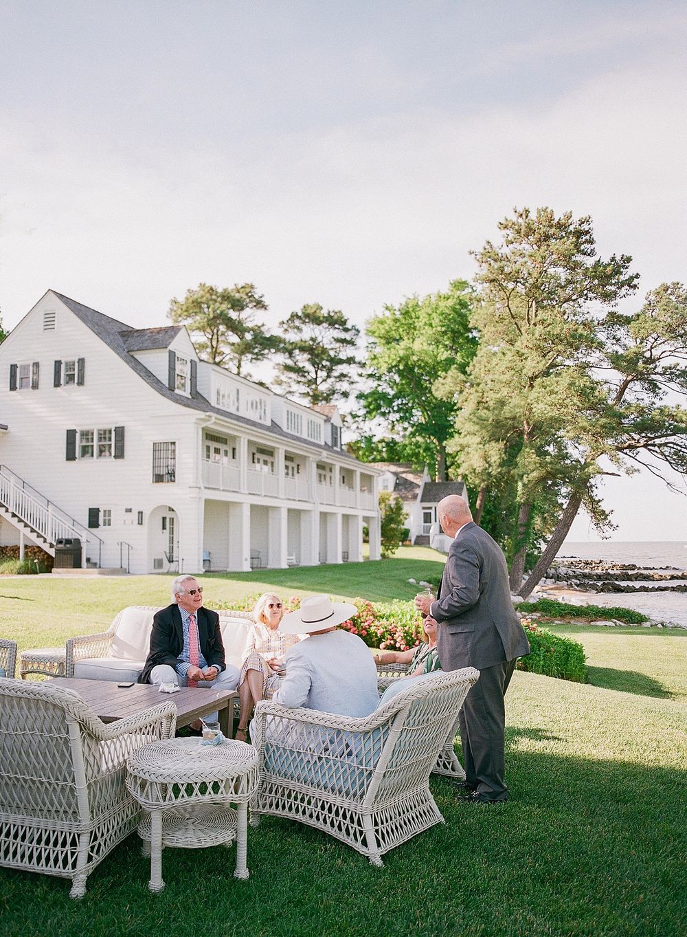 Elegant waterfront gathering with wicker furniture on a manicured lawn beside a white coastal house with garage doors.