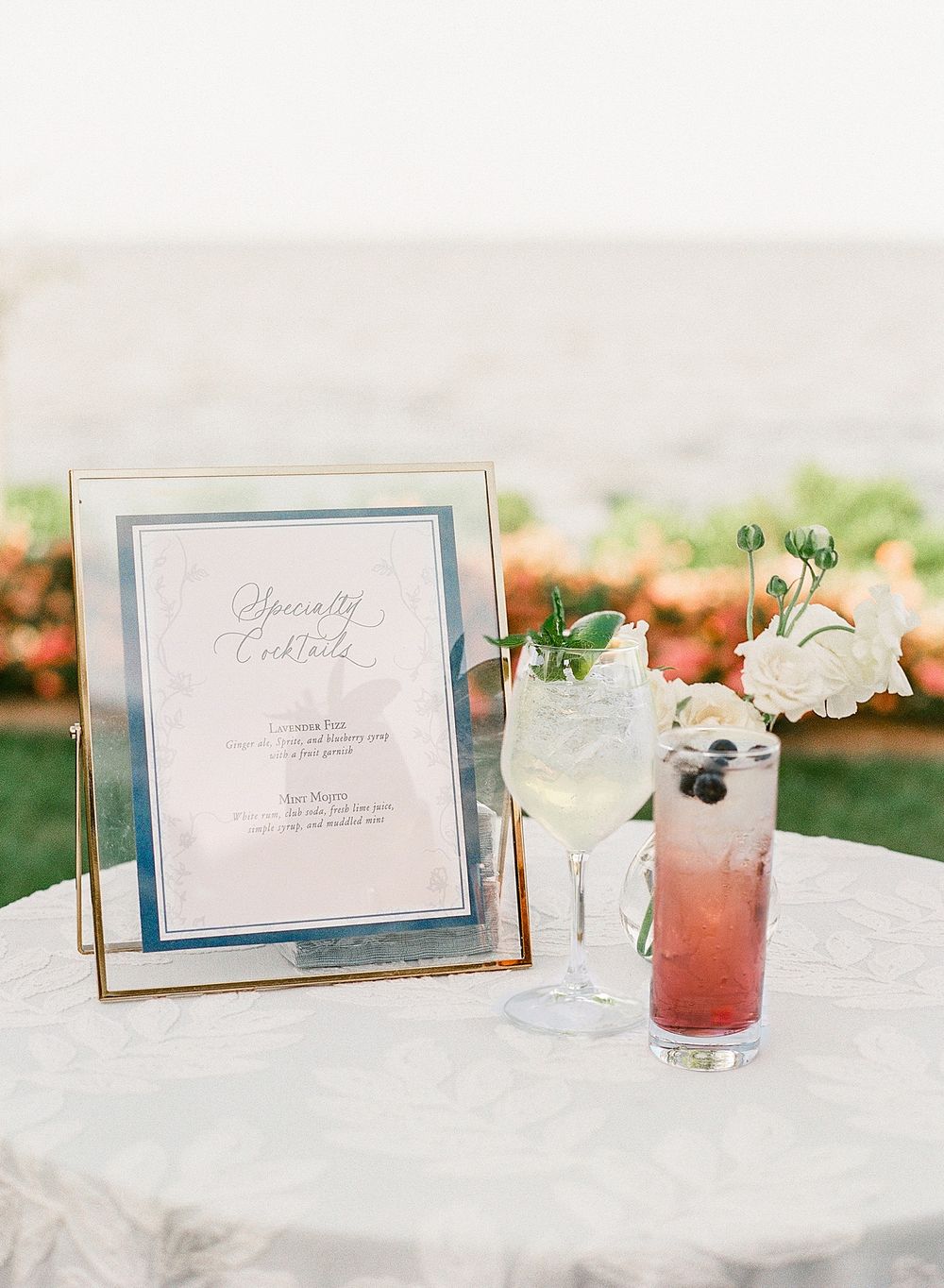 Elegant framed menu card displayed alongside colorful cocktails with floral garnishes on an outdoor table.
