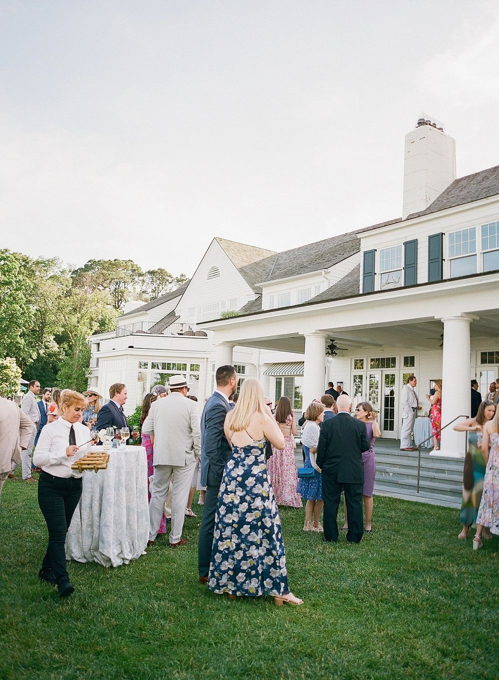 Guests enjoying an elegant outdoor reception at a white colonial-style house with black shutters on a sunny afternoon.