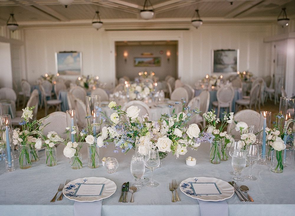 Elegant wedding reception table with white floral centerpieces, glass vases and ivory chair covers in a bright banquet room.