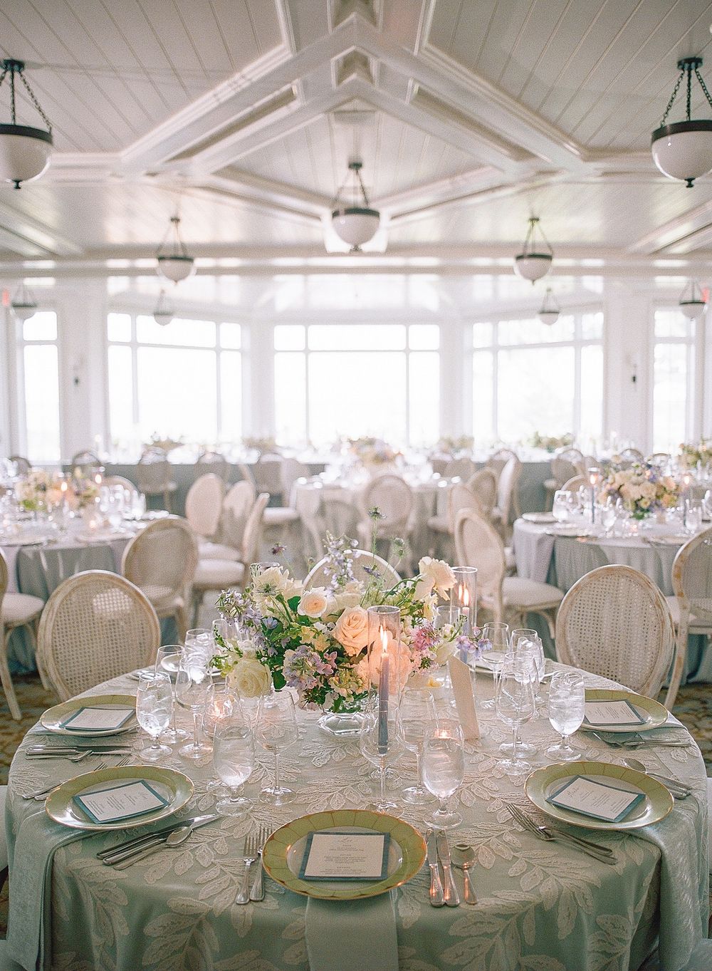 Elegant wedding reception ballroom with sage green tablecloths, vintage chairs and peach floral centerpieces under crystal chandeliers.