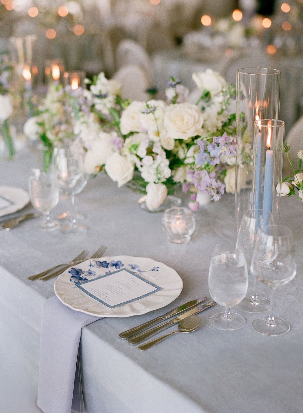 Elegant wedding table setting with white and purple floral centerpiece, taper candles, and blue-detailed place settings.