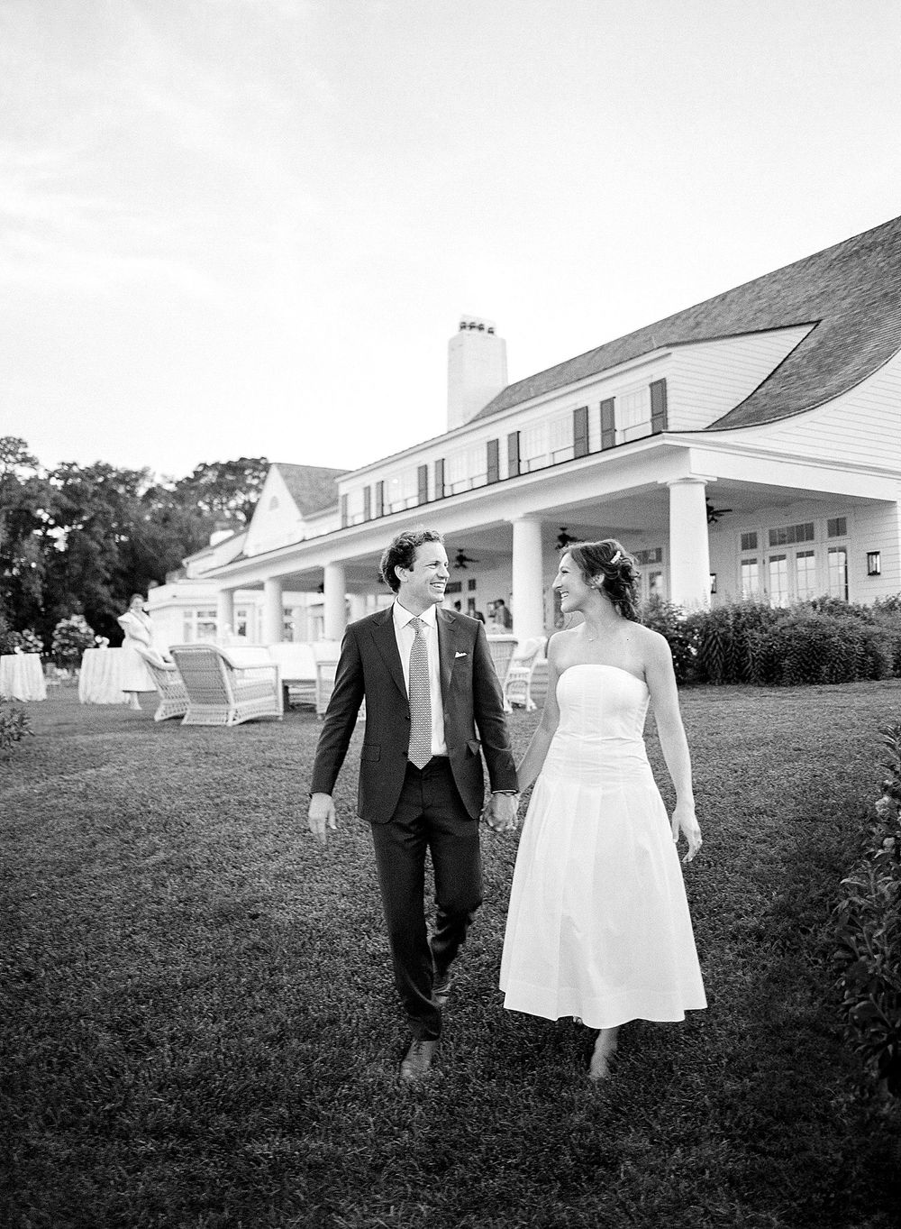 A couple in wedding attire walks together on the lawn of an elegant white house with a columned porch.