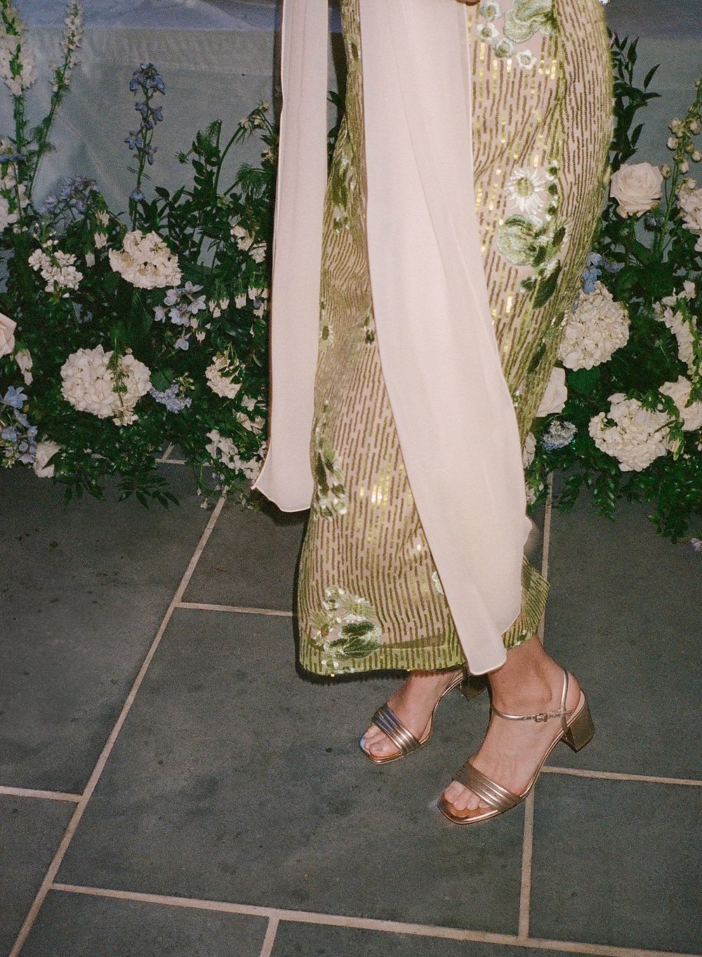 A person wearing a gold sequined dress and strappy sandals stands near white floral arrangements on tiled floor.