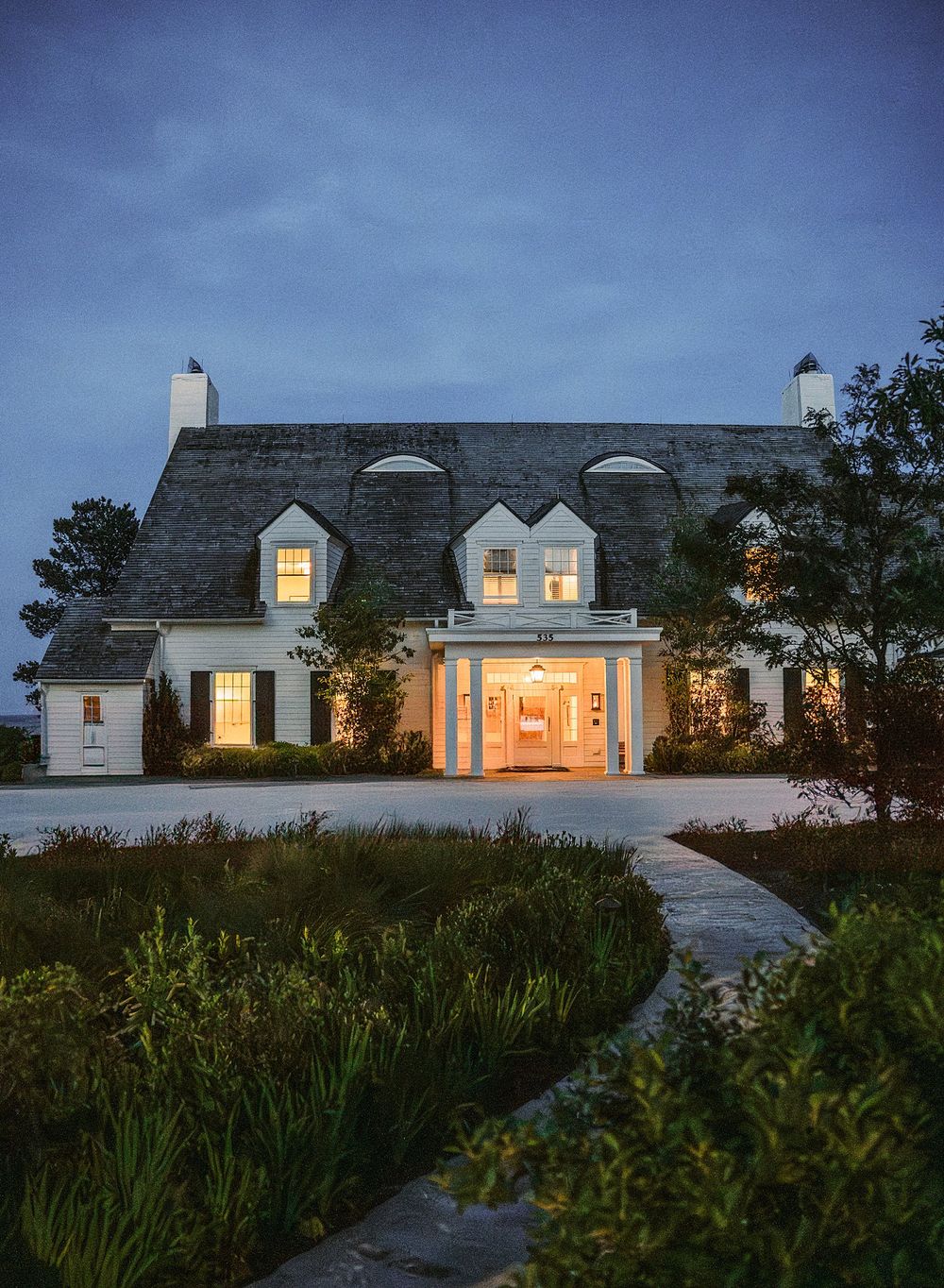 Elegant white cottage-style home with dormer windows and warm lighting glowing from within at dusk.