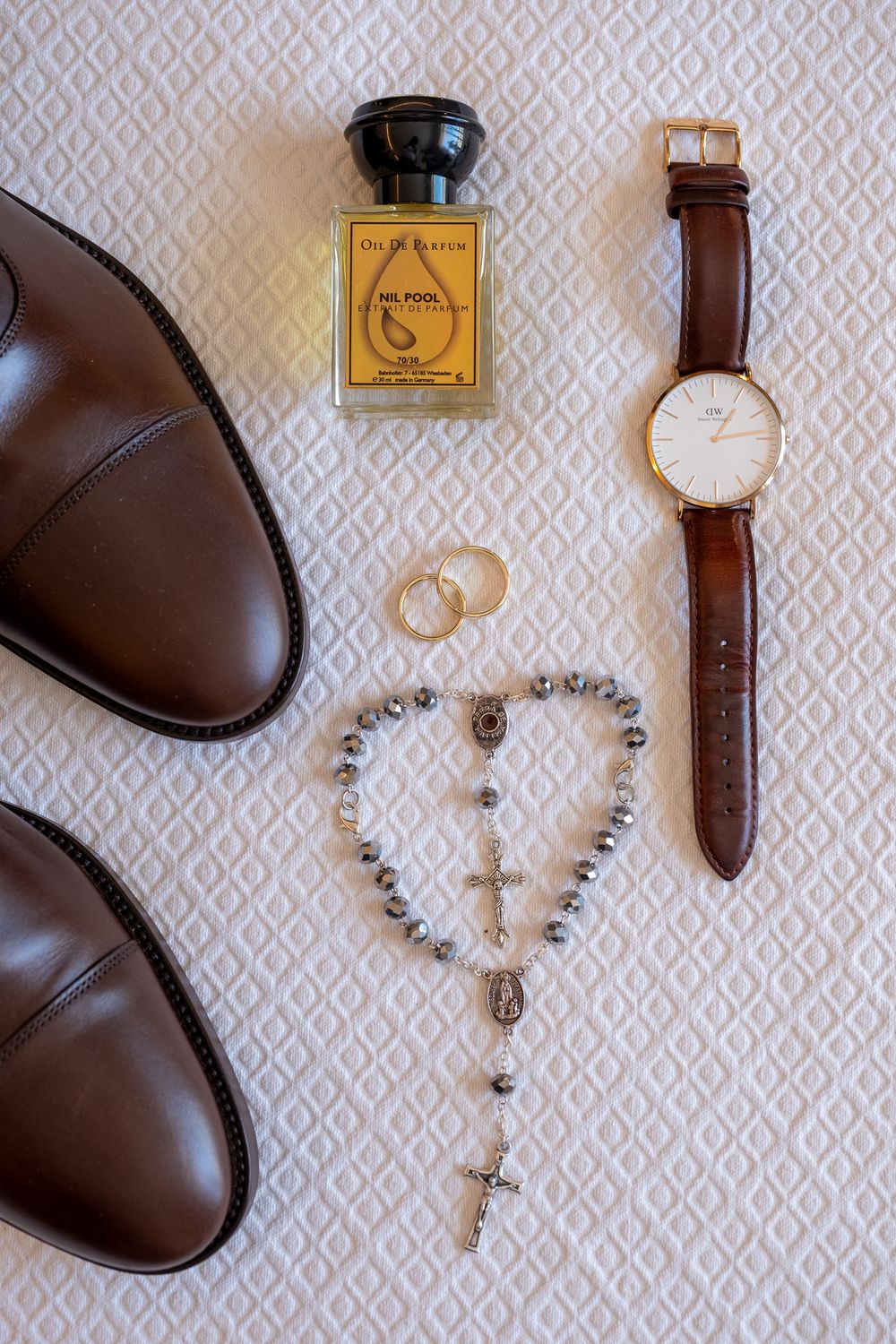 Wedding accessories including brown shoes, perfume, rosary beads, rings and leather watch on textured surface.
