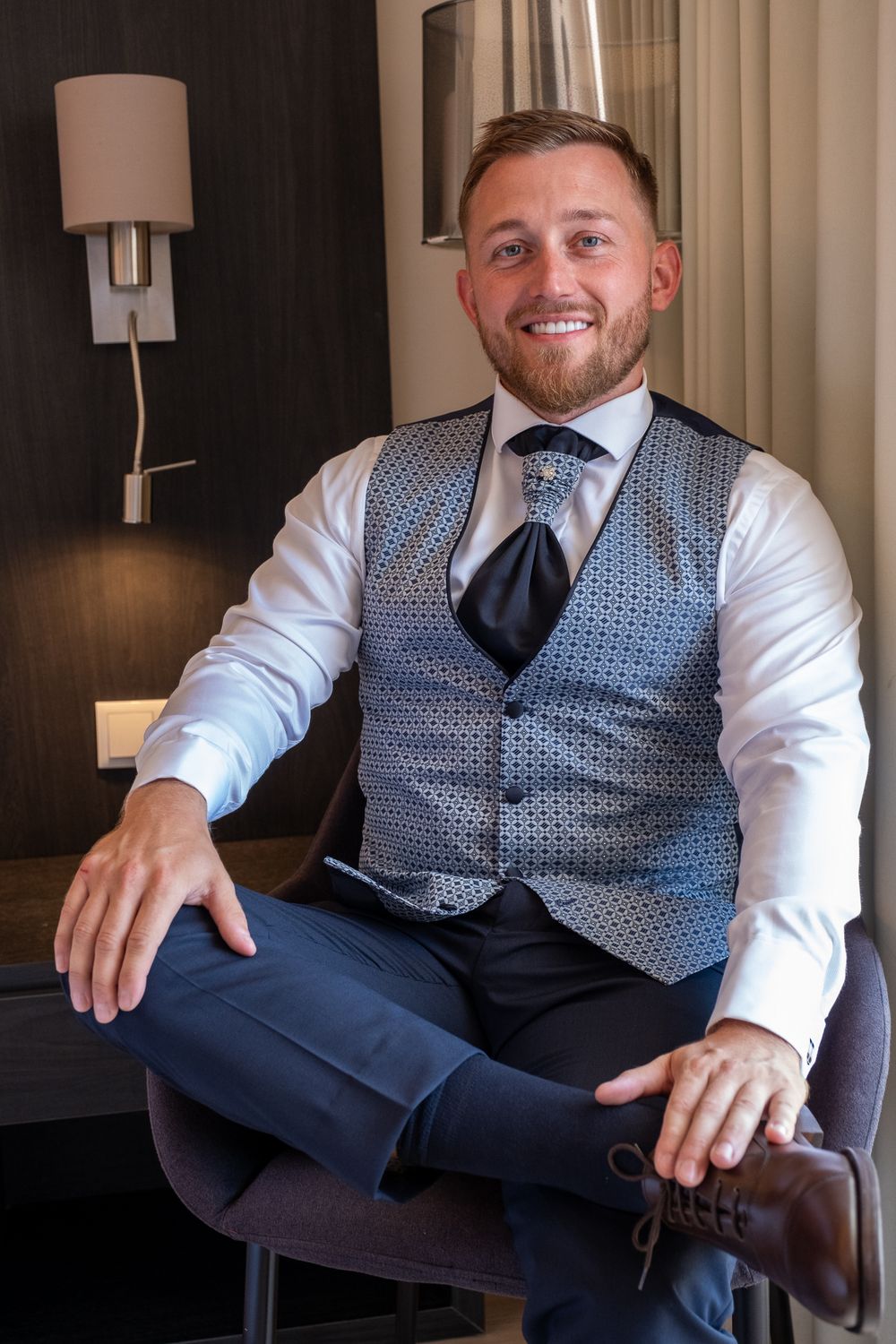 Wedding attire featuring patterned vest and tie while seated in hotel room.