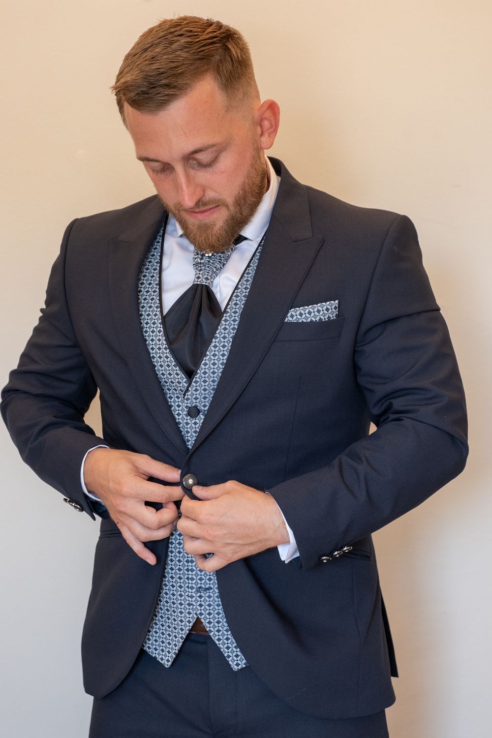 Man adjusts buttons on dark suit with light patterned vest and tie for formal occasion.