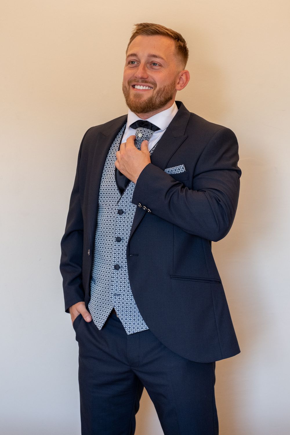 Sharp dressed figure adjusts blue tie while wearing modern navy suit with patterned vest.