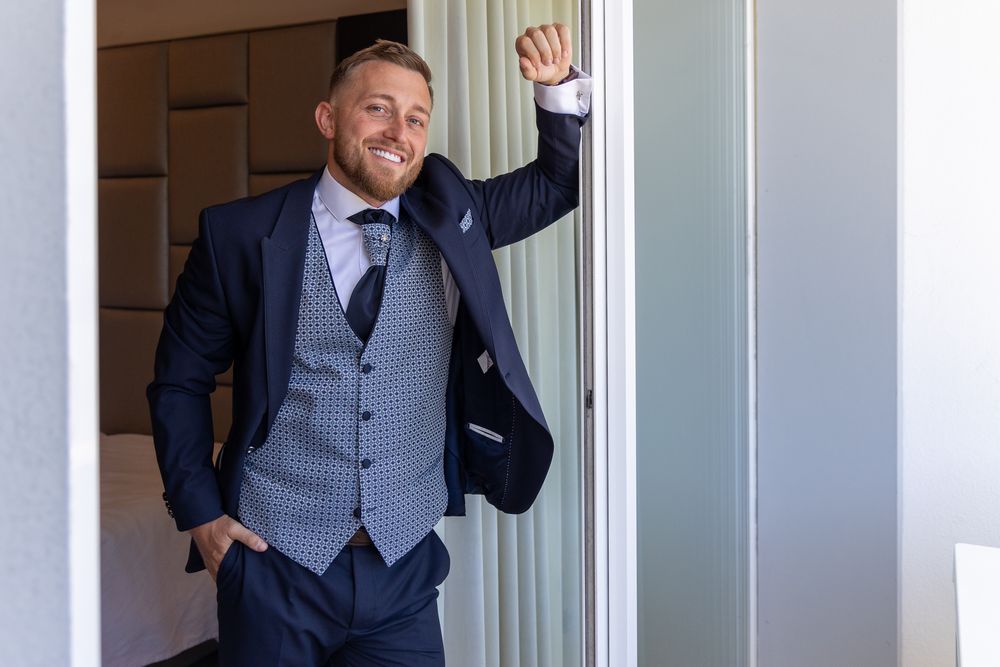 Figure waves from doorway while wearing tailored navy suit with checkered waistcoat.