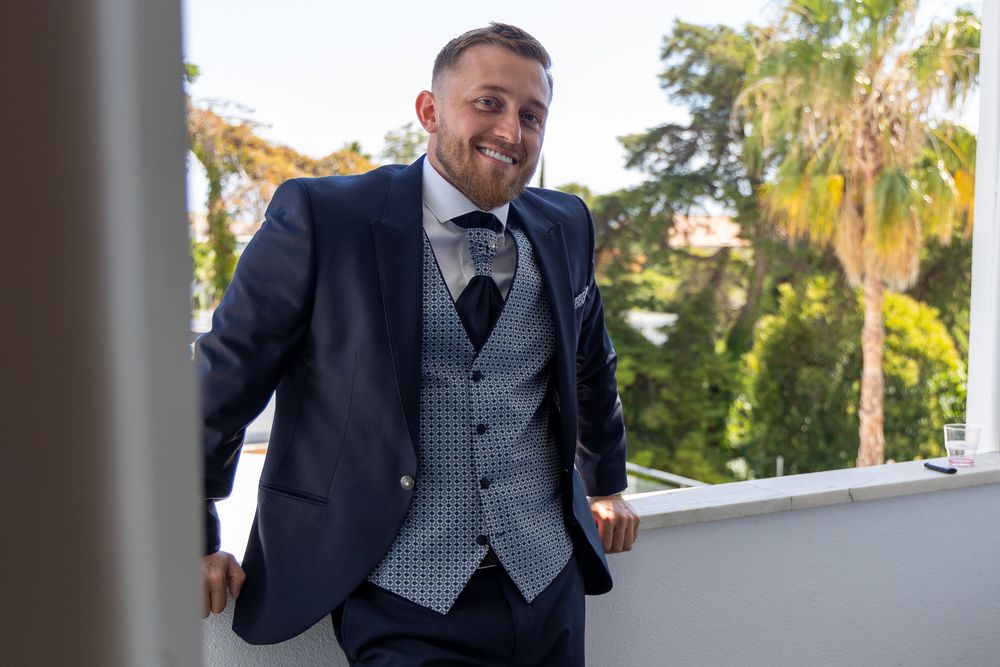 Suited figure stands on balcony with grey waistcoat and patterned tie.
