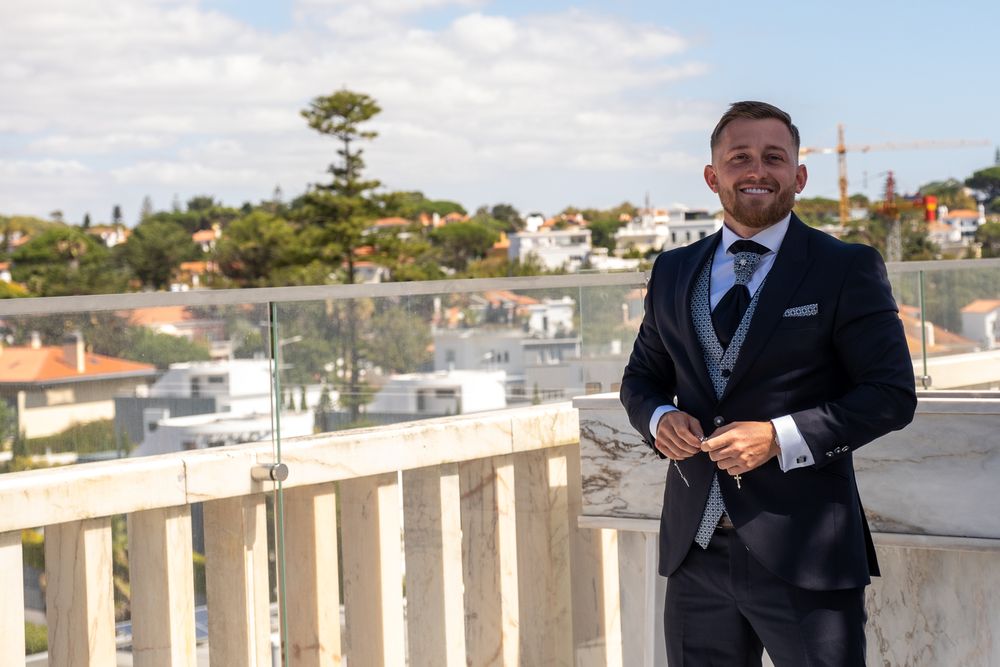 Suited figure stands on balcony overlooking coastal town with blue sky background.