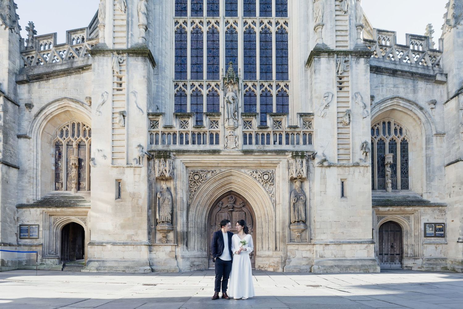 A couple stands in formal attire in front of an ornate Gothic cathedral entrance with stone archways and stained glass windows.