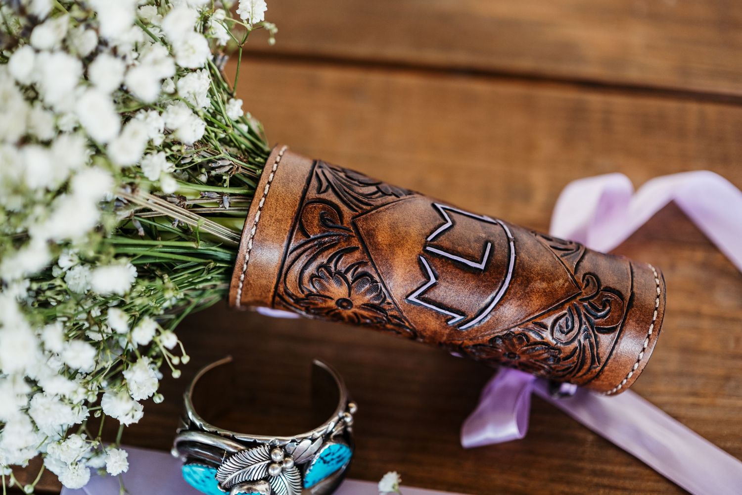 A carved leather drink sleeve with intricate designs sits beside delicate white baby's breath flowers and purple ribbon.