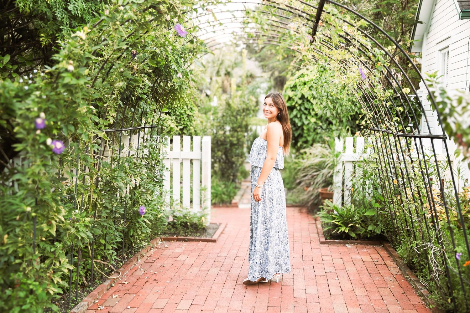 A figure stands beneath a garden arbor covered in vines on a brick pathway surrounded by flowers and greenery.