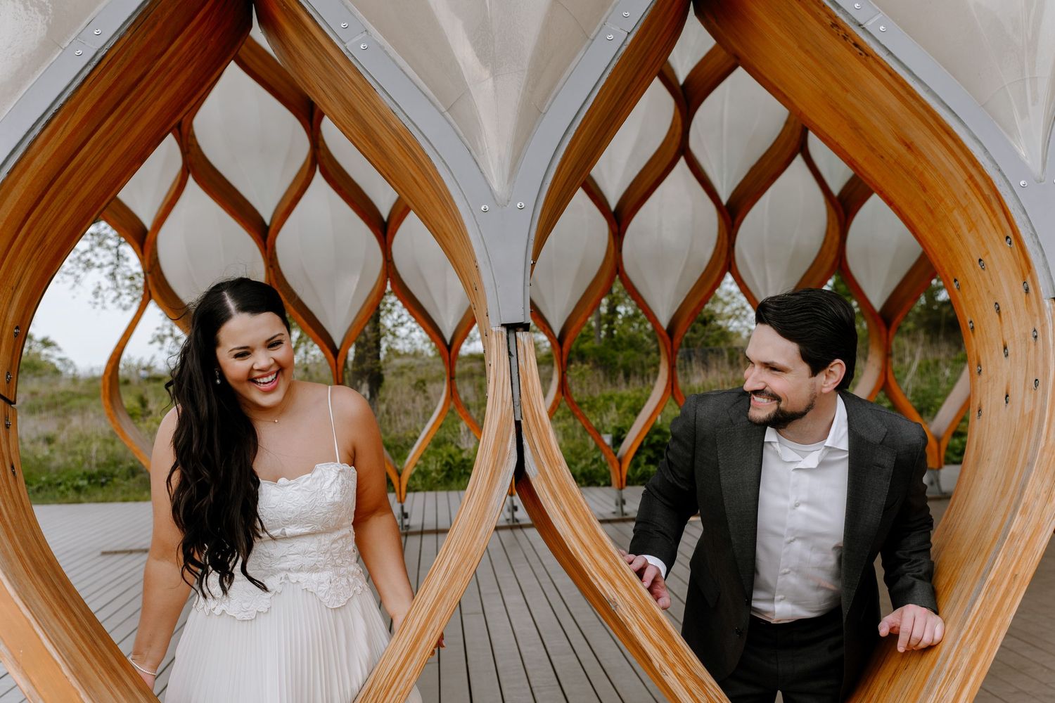 A couple in wedding attire shares a joyful moment through a decorative wooden archway on an outdoor deck.