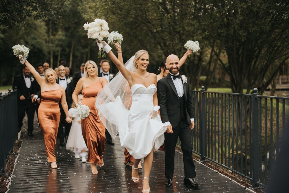 Wedding party runs joyfully down a wet boardwalk path while holding white bouquets in the rain at Grand Lodge Oaklands.