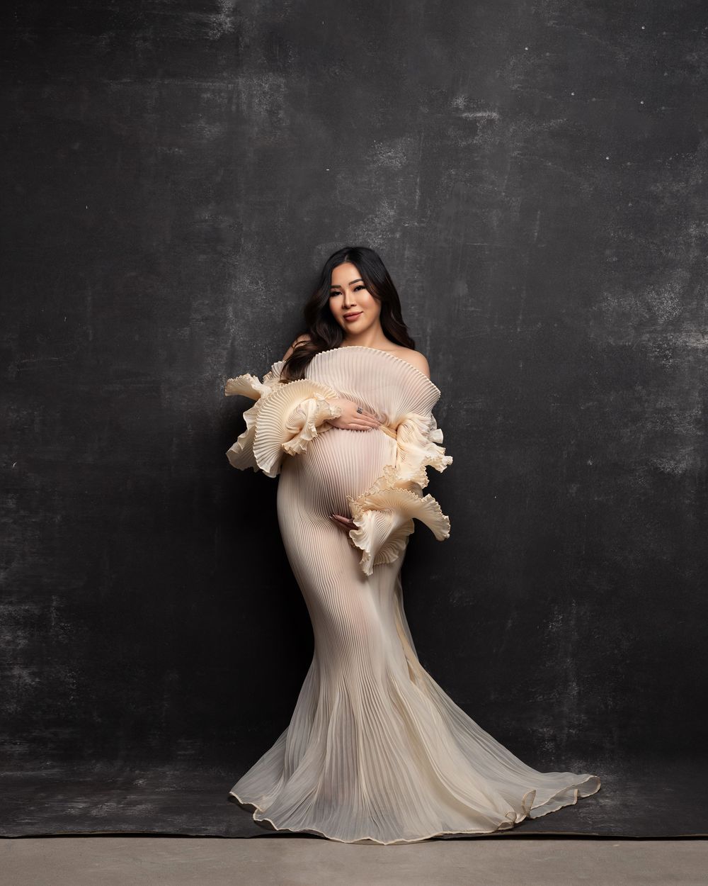 Elegant maternity portrait in a flowing cream-colored gown against a dark textured backdrop.