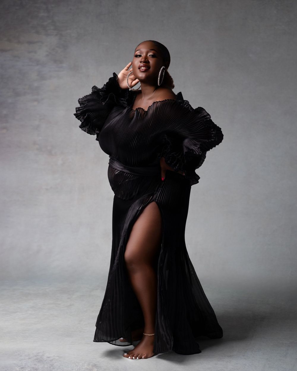 Glamorous figure in a flowing black evening gown with dramatic ruffled sleeves poses barefoot against grey studio backdrop.