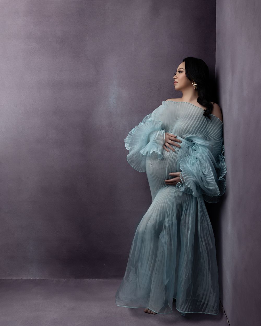 Elegant maternity photo featuring a flowy powder blue pleated gown with dramatic feathered bell sleeves against a dark backdrop.