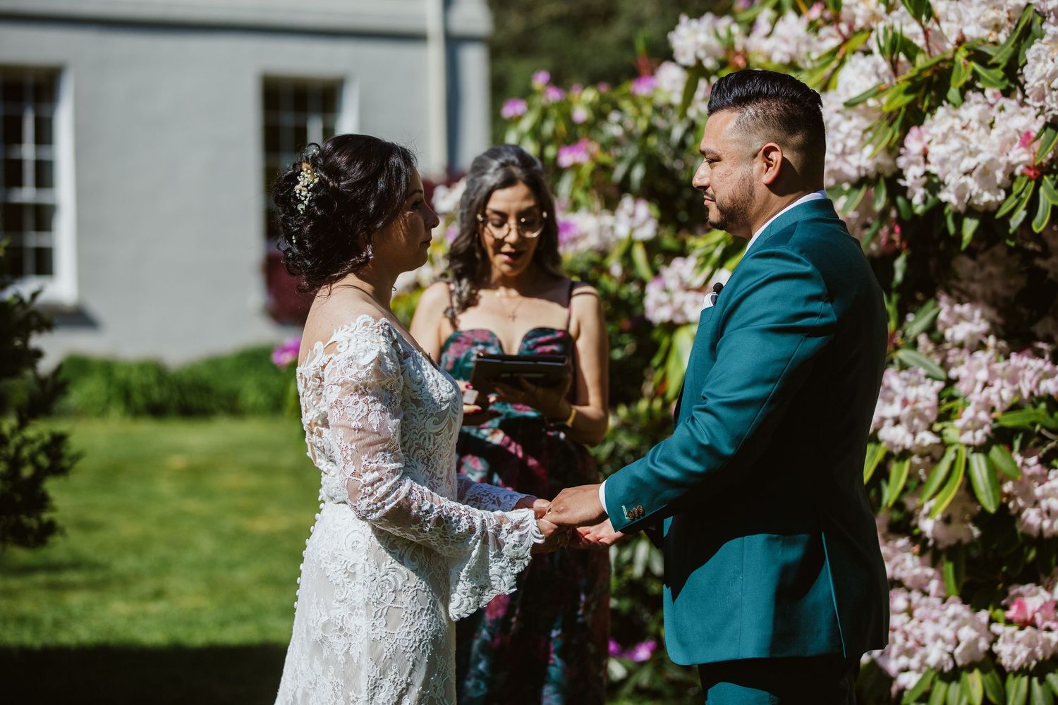 A couple celebrates their outdoor wedding ceremony with an officiant near blooming pink flowers in a garden setting.