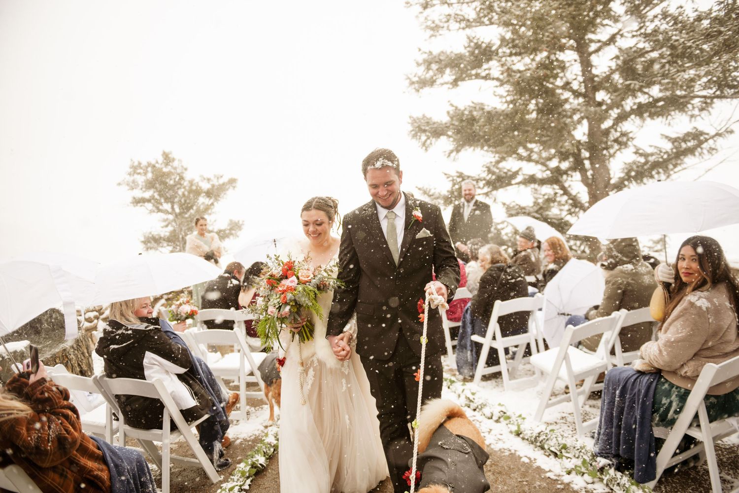 Wedding ceremony celebrations underway at an outdoor venue with guests seated on white chairs and autumn trees in background.