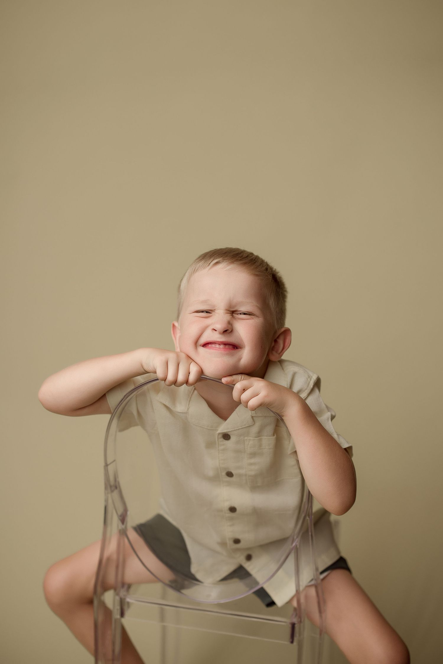 Young child sitting backwards on a clear chair wearing a light colored vest smiles joyfully against beige background.
