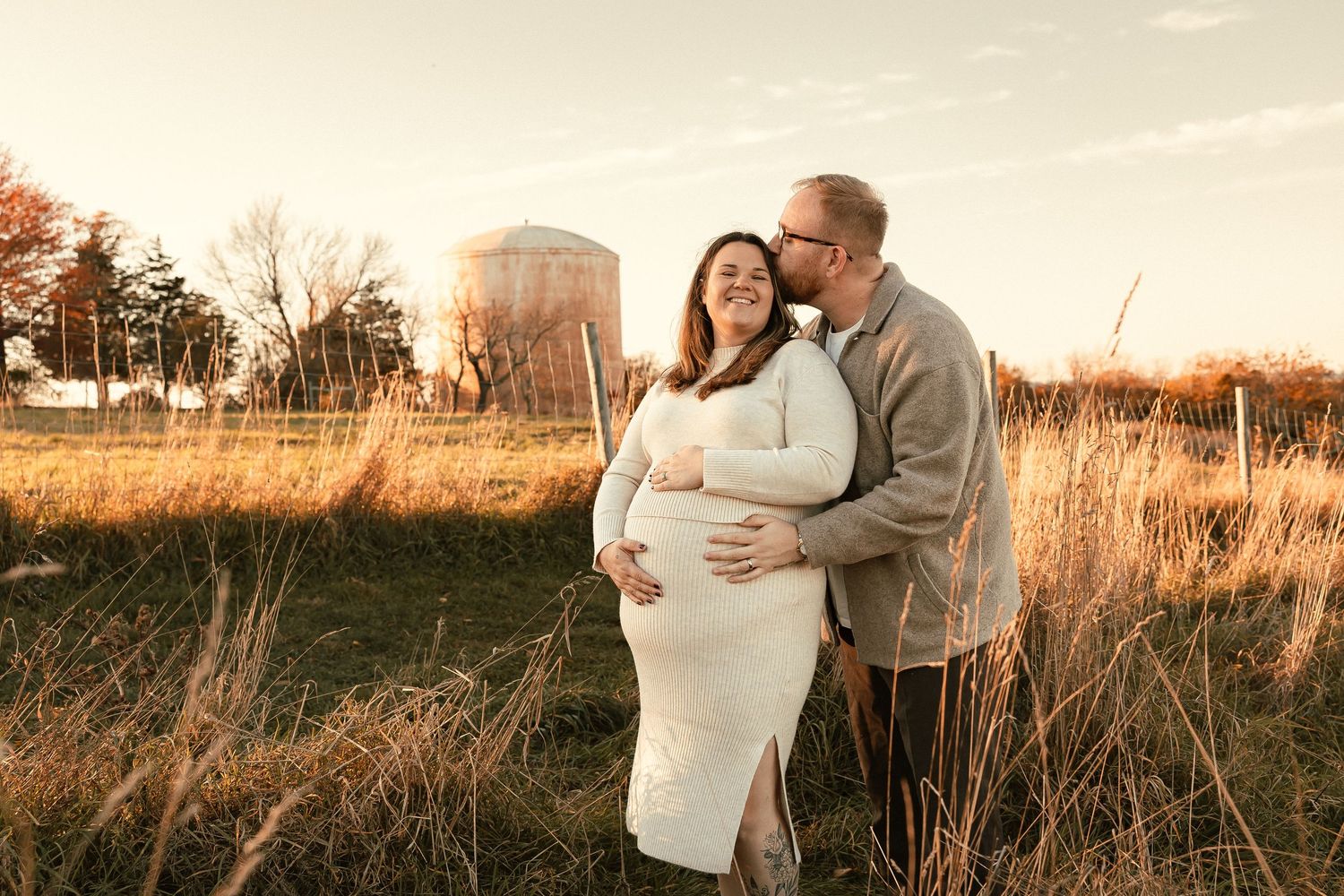 A couple embraces in a tall grass field during golden hour sunset casting a warm glow over their maternity photo session.