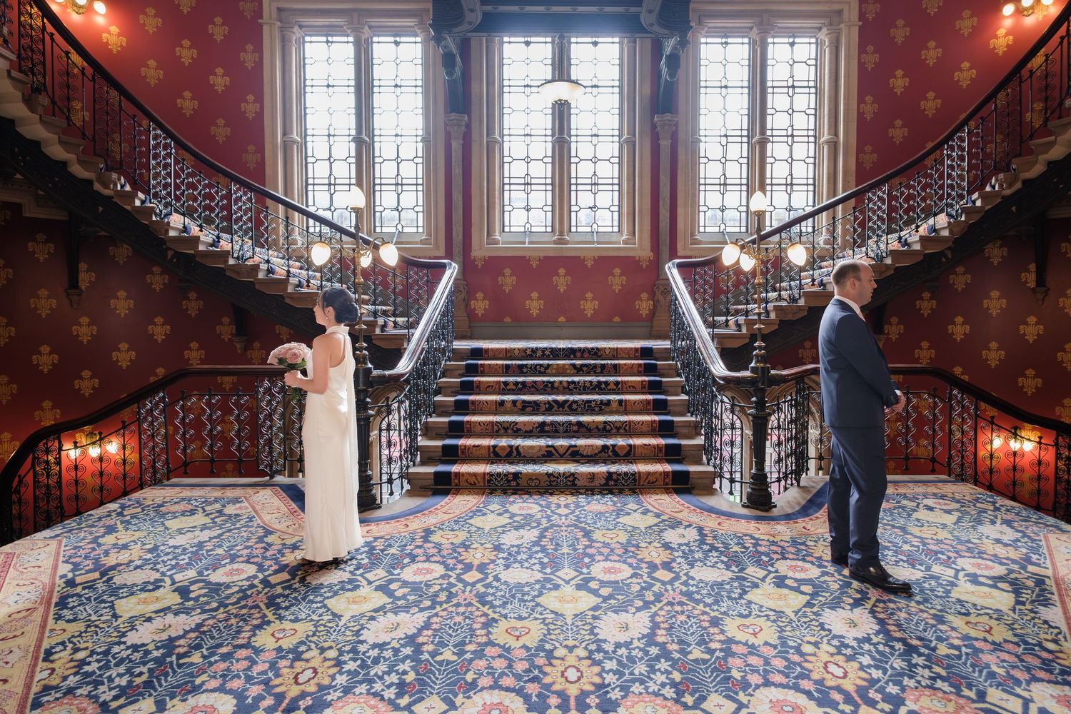 Two people stand on opposite sides of an elegant staircase with ornate carpet and red wallpaper in a historic building.