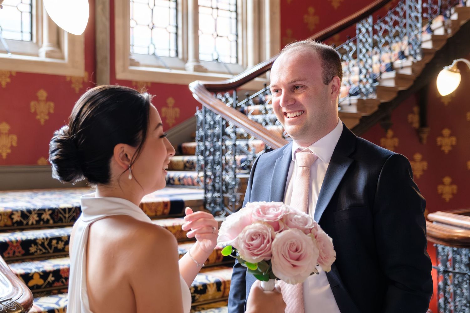 A couple shares a sweet moment near an ornate staircase, with pink roses and stained glass windows in the backdrop.