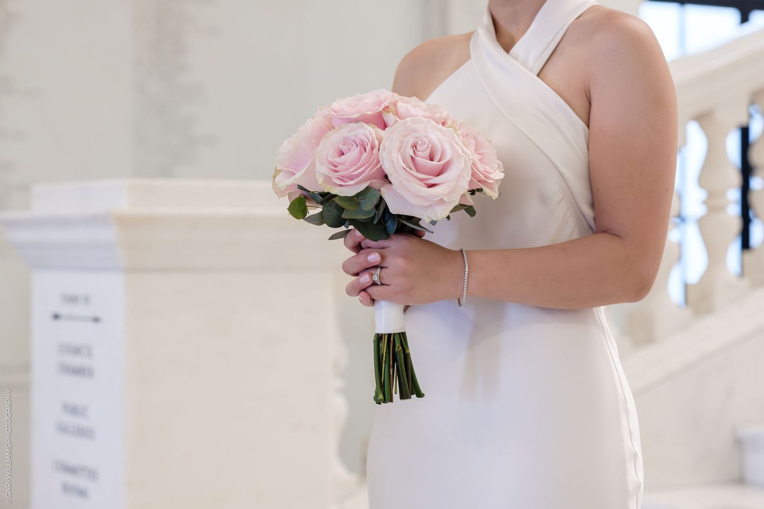 A bride in a classic white wedding dress holds a delicate pink rose bouquet in a sunlit room.