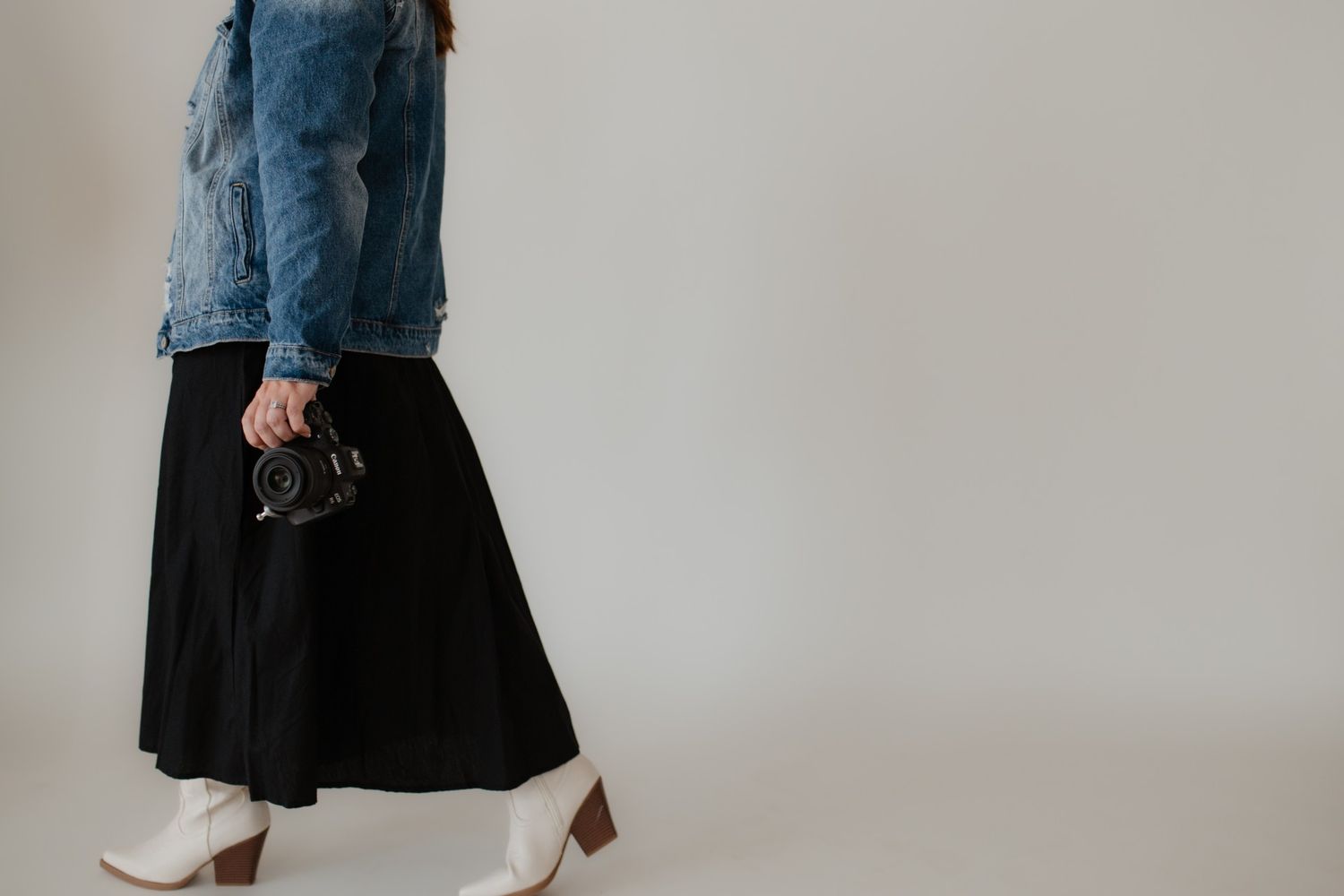 Person wearing a denim jacket and long black skirt walks against a white wall.