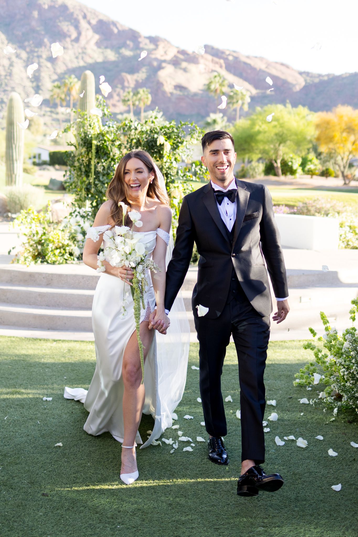A couple runs playfully through scattered flower petals on a grassy lawn with mountains in the background.