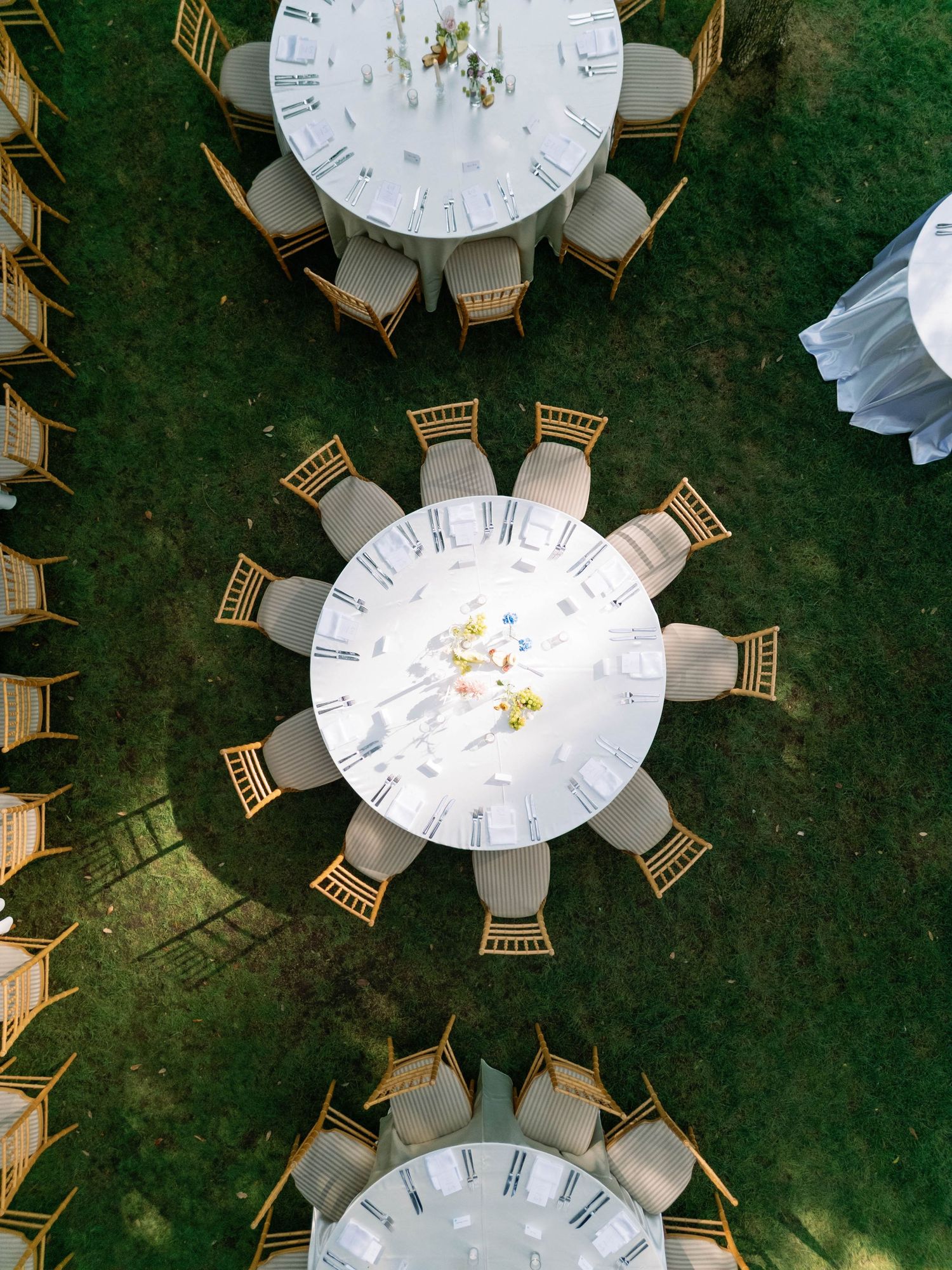 Aerial view of three elegant round white dining tables with gold chairs arranged on a green lawn for an outdoor event.