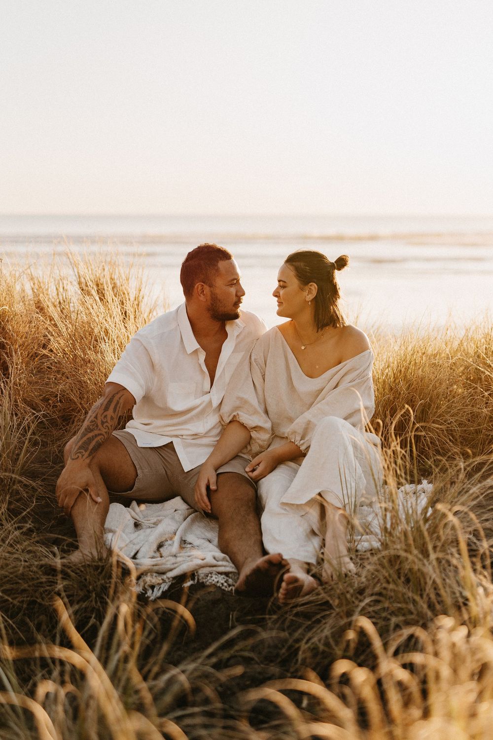 Couple in white clothes sitting together among tall grass at sunset on a beach.